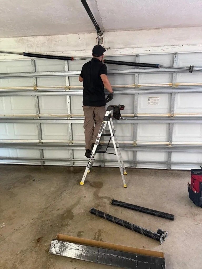Technician on a stepladder reaching a torsion tube above a white door, spring parts on the floor