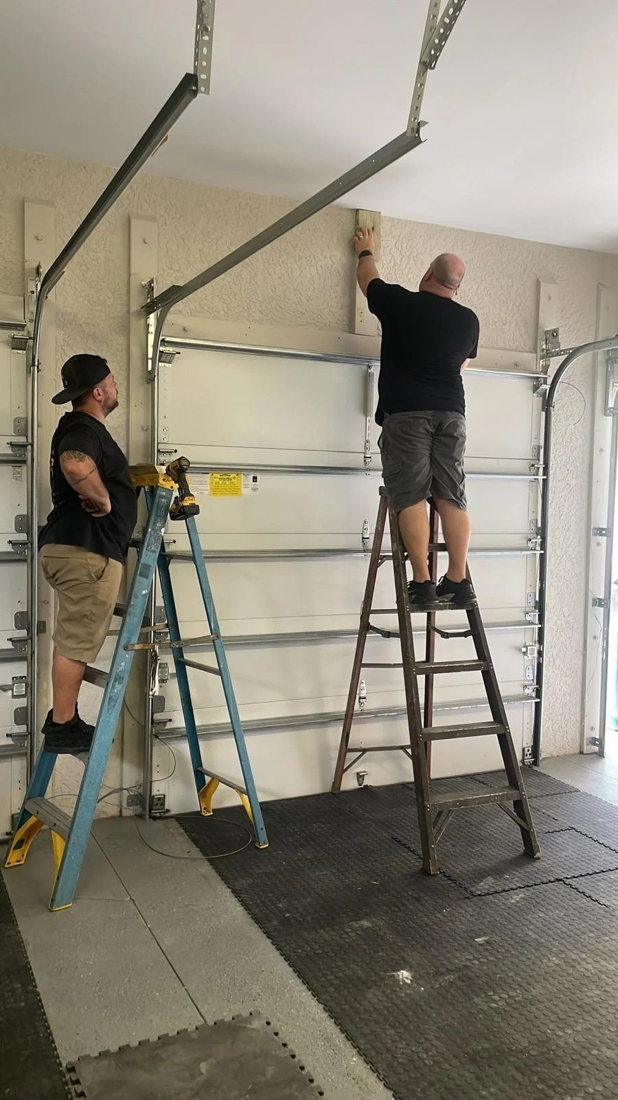 Two technicians on ladders working at the top of a white sectional door