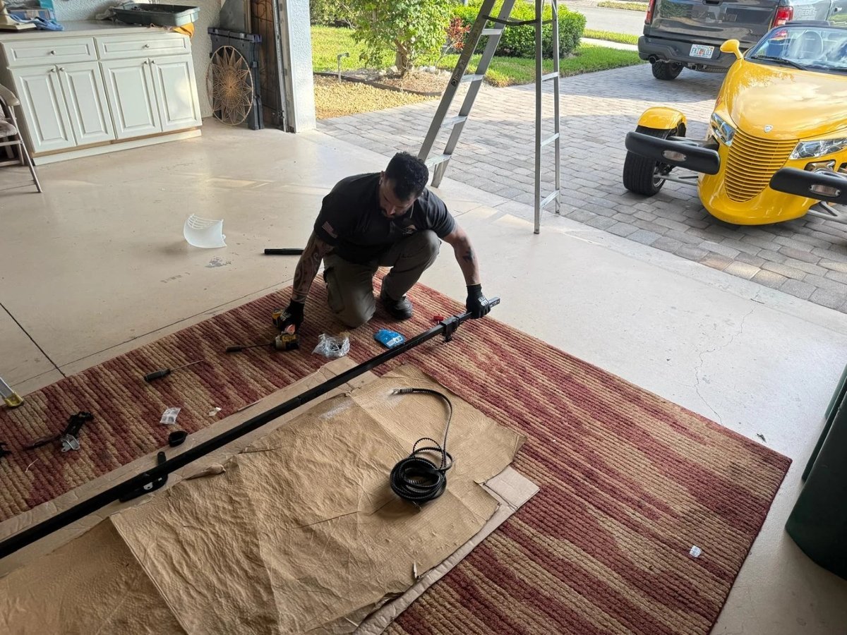 Technician kneeling on a rug beside a long metal garage door part, with a yellow car parked outside